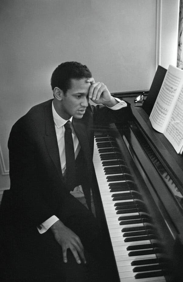 A black-and-white photo of Andre Watts, short-haired and wearing a suit and tie, sitting at a piano with his head resting on his left hand.