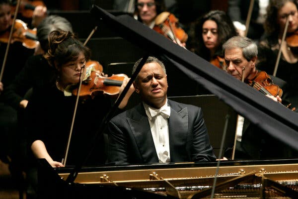 Mr. Watts, formally dressed, sits at a piano with an intense look on his face. Members of the New York Philharmonic are behind him.